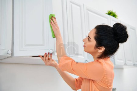A woman in casual attire meticulously cleans a kitchen using a green rag, ensuring every surface shines with a sparkle.