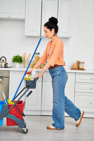 A stylish woman in casual attire pushes a stroller through a cluttered kitchen while multitasking at home.