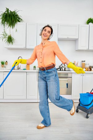 A stylish woman in casual attire gracefully mops the kitchen floor with a bucket nearby.