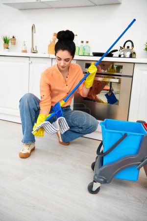 Une femme en tenue décontractée nettoie gracieusement le sol avec une serpillière et un seau dans sa maison.