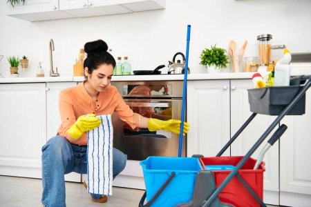 Una mujer elegante con atuendo casual limpiando el piso con una fregona y un cubo en casa.