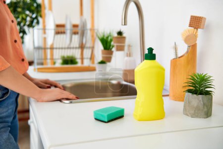 A stylish woman in casual attire stands in a kitchen next to a sink, cleaning and tidying up the space.