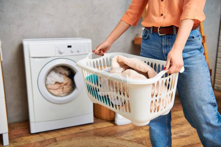 A stylish woman in casual attire holds a laundry basket in front of a washing machine.