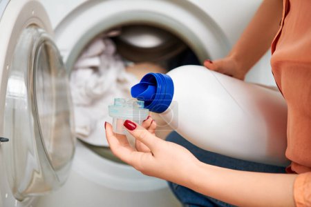 Eine stilvolle Frau hält eine Flasche Wasser vor einer Waschmaschine, während sie ihr Haus putzt.