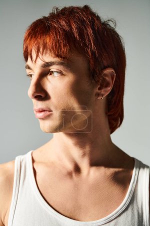 Stylischer junger Mann mit roten Haaren posiert selbstbewusst in einem Tank-Top vor grauem Studiohintergrund.