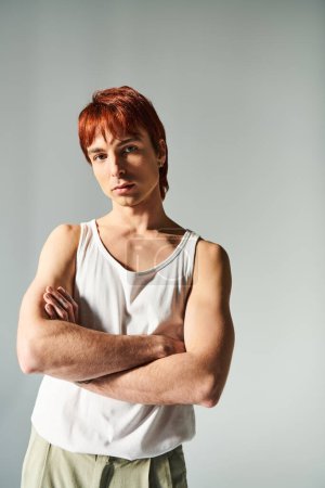 Foto de A stylish young man with red hair stands confidently with his arms crossed in a studio against a grey background. - Imagen libre de derechos