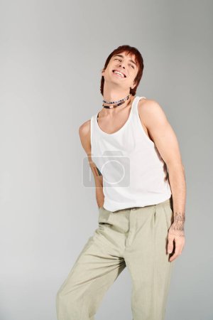 Photo for A stylish young man poses confidently in a studio setting, wearing a white tank top and khaki pants against a grey background. - Royalty Free Image