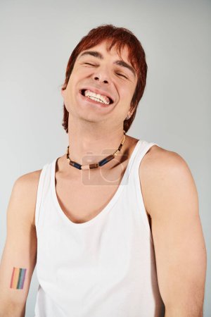 Photo for A fashionable young man strikes a pose in a white tank top, accessorized with a vibrant rainbow necklace, against a grey studio backdrop. - Royalty Free Image
