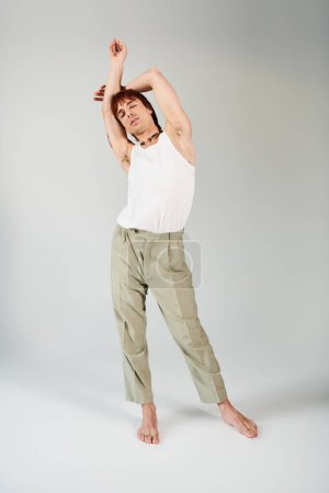Photo for A stylish young man posing confidently in a studio, wearing a white tank top and khaki pants, against a grey background. - Royalty Free Image