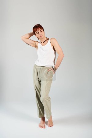 A stylish young man striking a pose in a white tank top and khaki pants against a grey studio background.