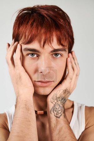 Ein stilvoller junger Mann mit roten Haaren hält sich vor grauem Hintergrund die Hände vor das Gesicht, tief in Gedanken.