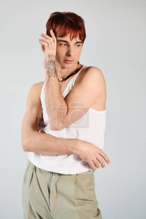 A young man strikes a pose in a studio, wearing a tank top and khaki pants against a grey background.