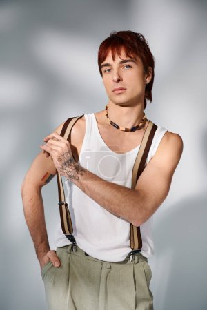 Foto de Un joven elegante con el pelo rojo posa con confianza en una camisa blanca y tirantes contra un fondo de estudio gris. - Imagen libre de derechos