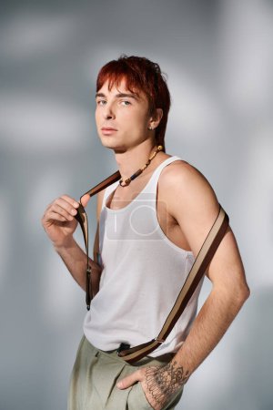 Téléchargez les photos : Un jeune homme élégant avec les cheveux roux frappant une pose dans un débardeur blanc sur un fond de studio gris. - en image libre de droit