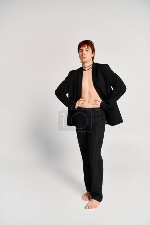 A stylish young man in a suit confidently stands with his hands on his hips in a studio with a grey background.