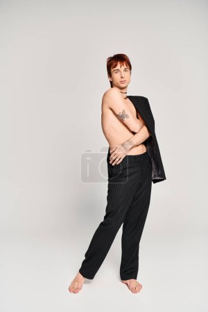 Téléchargez les photos : A stylish young man with a sculpted physique poses in black pants against a grey background in a studio setting. - en image libre de droit