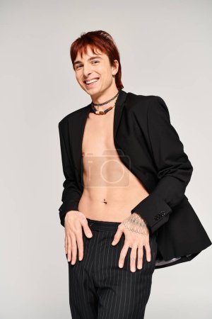 A stylish shirtless man in a suit strikes a pose in a studio setting with a grey background.