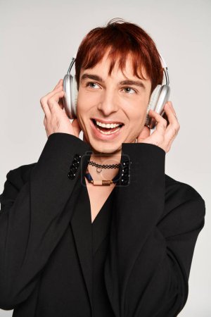 A stylish young man with vibrant red hair listens to music on headphones against a grey studio backdrop.