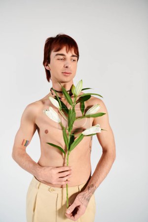 Téléchargez les photos : Un homme torse nu tient tendrement une plante dans ses mains, montrant un lien avec la nature dans un cadre de studio sur un fond gris. - en image libre de droit