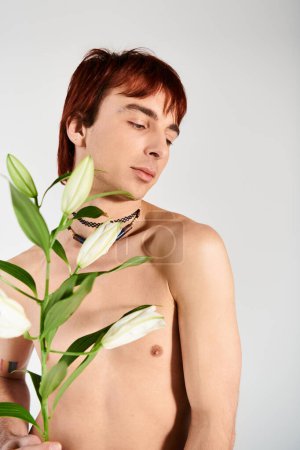 Hemdloser junger Mann mit einer zarten Blume in einem Atelier vor grauem Hintergrund, die ein Gefühl von Ruhe und Schönheit ausstrahlt.