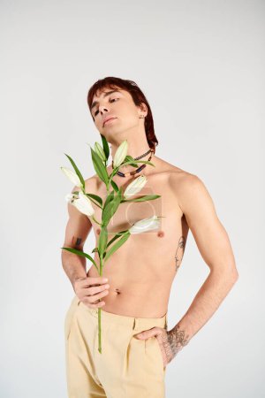 Ein junger Mann hält selbstbewusst eine zarte Blume in einem Atelier vor grauem Hintergrund und strahlt ein Gefühl von Intimität und Schönheit aus.
