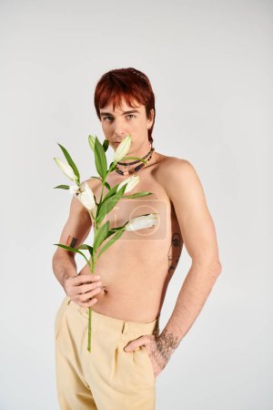 Téléchargez les photos : Un jeune homme torse nu pose sensuellement avec une fleur dans la bouche dans un décor de studio avec un fond gris. - en image libre de droit