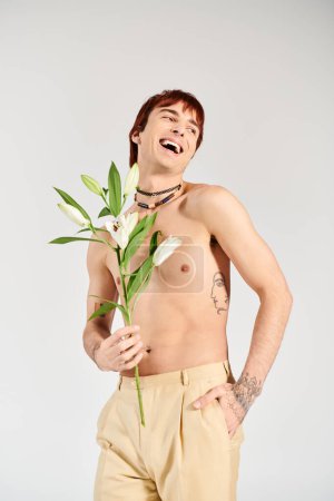 Ein junger Mann posiert selbstbewusst ohne Hemd und hält eine zarte Blume in einem Atelier vor grauem Hintergrund.