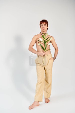 Photo for A young man poses with flowers in a studio, standing with hand in pocket in front of a grey background. - Royalty Free Image