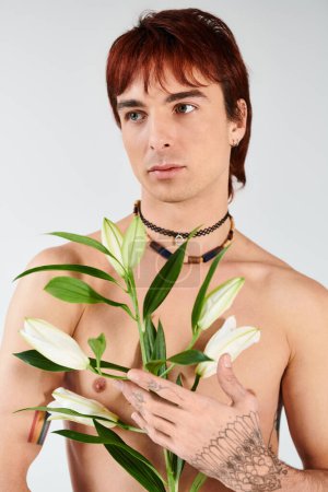 Foto de A shirtless young man in a studio holds a vibrant plant in his hand against a grey background. - Imagen libre de derechos
