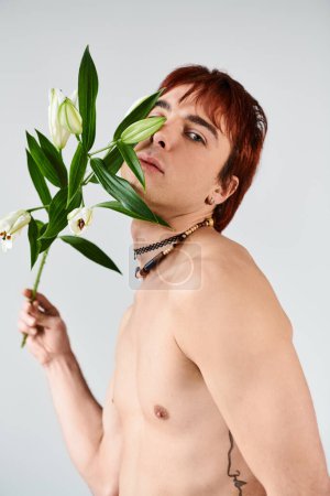 A young, shirtless man peacefully holds a delicate flower in a studio setting with a grey background.
