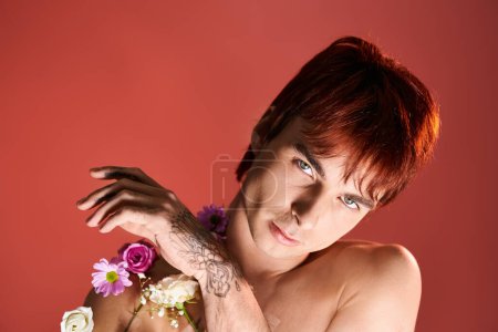 Téléchargez les photos : Un jeune homme se tient torse nu, berçant des fleurs vibrantes sur fond rouge. - en image libre de droit