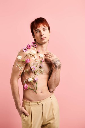 Ein junger Mann ohne Hemd posiert selbstbewusst mit Blumen, die seine Brust in einem Studio mit rosa Hintergrund schmücken.