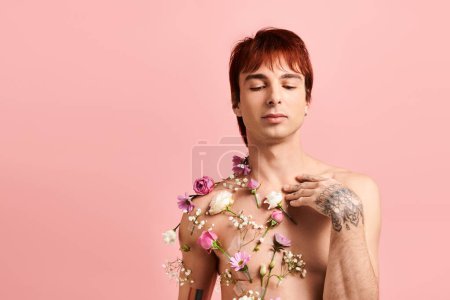 Ein junger Mann zeigt in einem Studio mit rosa Hintergrund stolz aufwändige Tätowierungen auf seiner Brust, die mit leuchtenden Blumen geschmückt sind.
