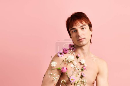 A shirtless young man poses confidently with vibrant flowers adorning his chest in a studio setting with a pink background