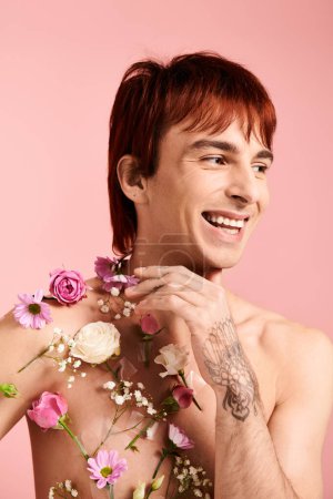 A shirtless young man poses with vibrant flowers adorning his chest in a studio against a pink background.