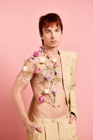 A shirtless young man confidently poses with colorful flowers adorning his chest against a pink studio backdrop.