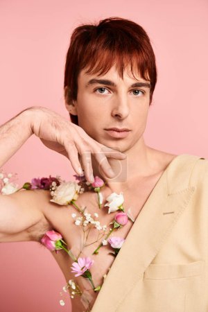 Un jeune homme aux cheveux roux pose, des fleurs ornant sa poitrine nue, sur fond de studio rose.