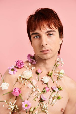 Ein junger Mann ohne Hemd posiert vor einem soliden rosafarbenen Hintergrund mit einer Vielzahl leuchtender Blumen, die seine Brust zieren..