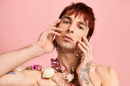 A young man with chest tattoos poses with flowers in a studio against a pink background.