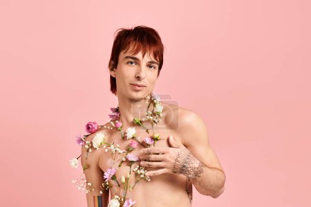Hemdloser Mann, der mit um den Hals drapierten Blumen in einem Atelier mit rosa Hintergrund posiert.