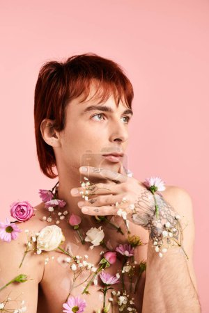 A shirtless young man strikes a pose with colorful flowers spread across his chest, set against a pink studio background.