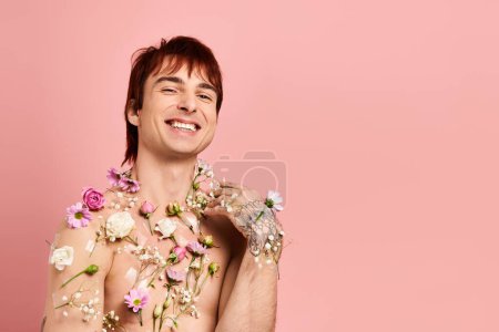 Un joven sin camisa posa delicadamente con flores vibrantes adornando su pecho sobre un fondo rosa.