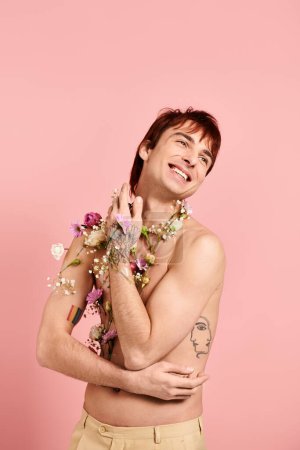 Téléchargez les photos : Un jeune homme, torse nu, exhibant fièrement des fleurs sur sa poitrine dans un studio avec un fond rose. - en image libre de droit