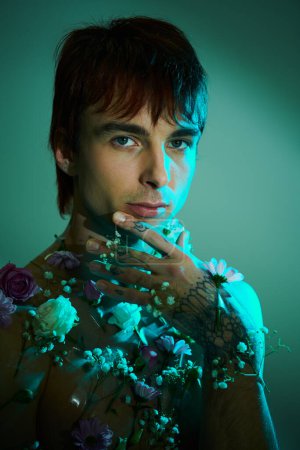 A young man confidently showcases his chest adorned with colorful tattoos and vibrant flowers in a studio setting against a blue background.