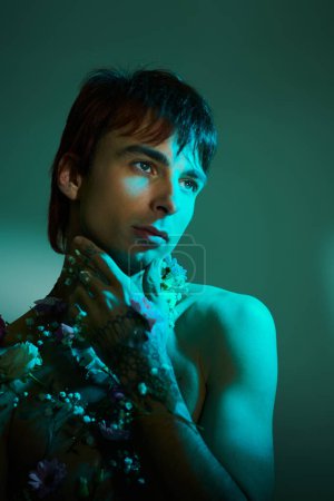 A stylish young man posing with flowers around his neck in a studio setting with a blue background.