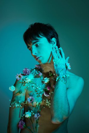 A stylish young man posing with flowers around his body in a studio setting with a blue light.
