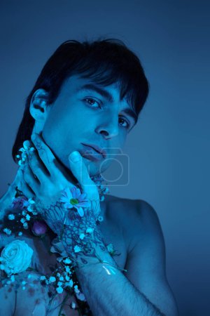 Ein junger Mann zeigt stolz seine aufwändigen Tätowierungen auf Armen und Brust, während er in einem Atelier mit blauem Licht von Blumen umgeben ist