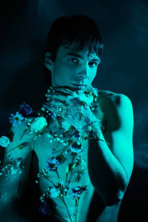 A young man confidently poses shirtless, with flowers delicately placed on his chest, against a blue light