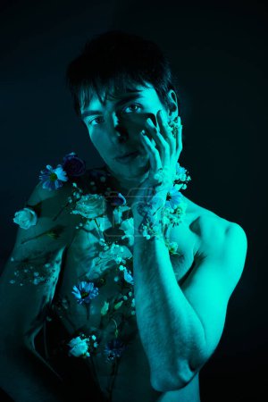Ein junger Mann ohne Hemd in einem von Blumen umgebenen Studio, das eine Mischung aus Männlichkeit und Weichheit zeigt.