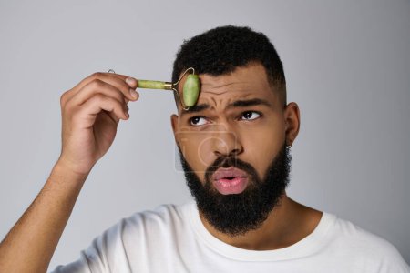 African american bearded man holds face roller for skincare routine.